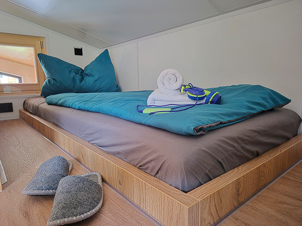 Ein einladendes Einzelbett im Alpenchalet Wendelstein mit Blick auf die unberührte Natur. Das Bett ist mit blaugrüner Bettwäsche bezogen; darauf liegen ein frisches weißes Handtuch und Kopfhörer. Im Vordergrund stehen graue handgemachte Filzpantoffeln auf dem hellen Echtholzboden im lichtdurchfluteten Obergeschoss eines alpinen modernem Ferienhauses.